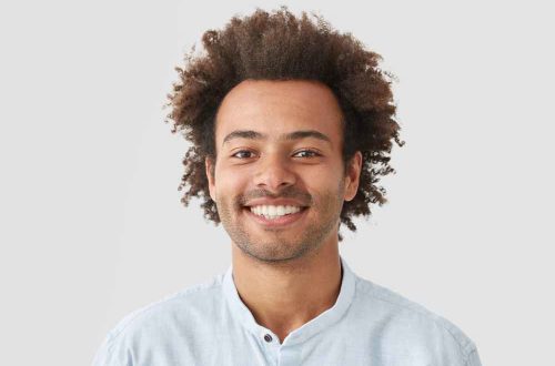 curly-man-with-broad-smile-shows-perfect-teeth-being-amused-by-interesting-talk-has-bushy-curly-dark-hair-stands-indoor-against-white-blank-wall